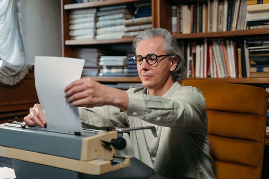 Senior man with glasses types on vintage typewriter in a library with bookshelves.