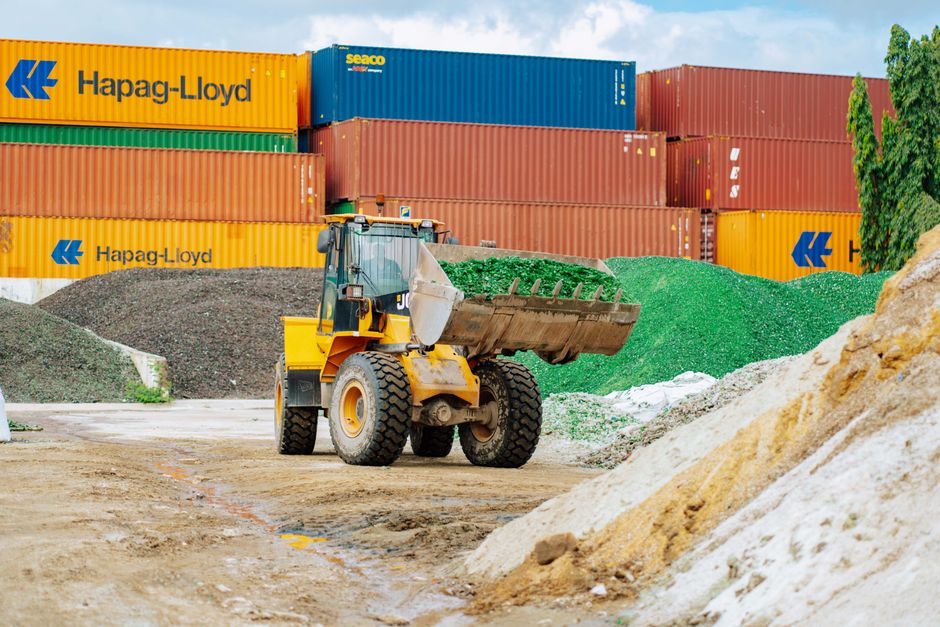 A bulldozer moves glass materials at a recycling yard with colorful shipping containers in Dar es Salaam.